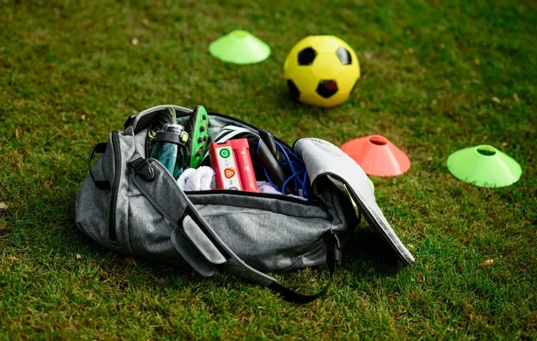 An open gray duffel bag packed with soccer cleats, a water bottle, a towel, and a red St John Pocket AED defibrillator, sitting on grass next to a yellow soccer ball and orange and green cone markers.