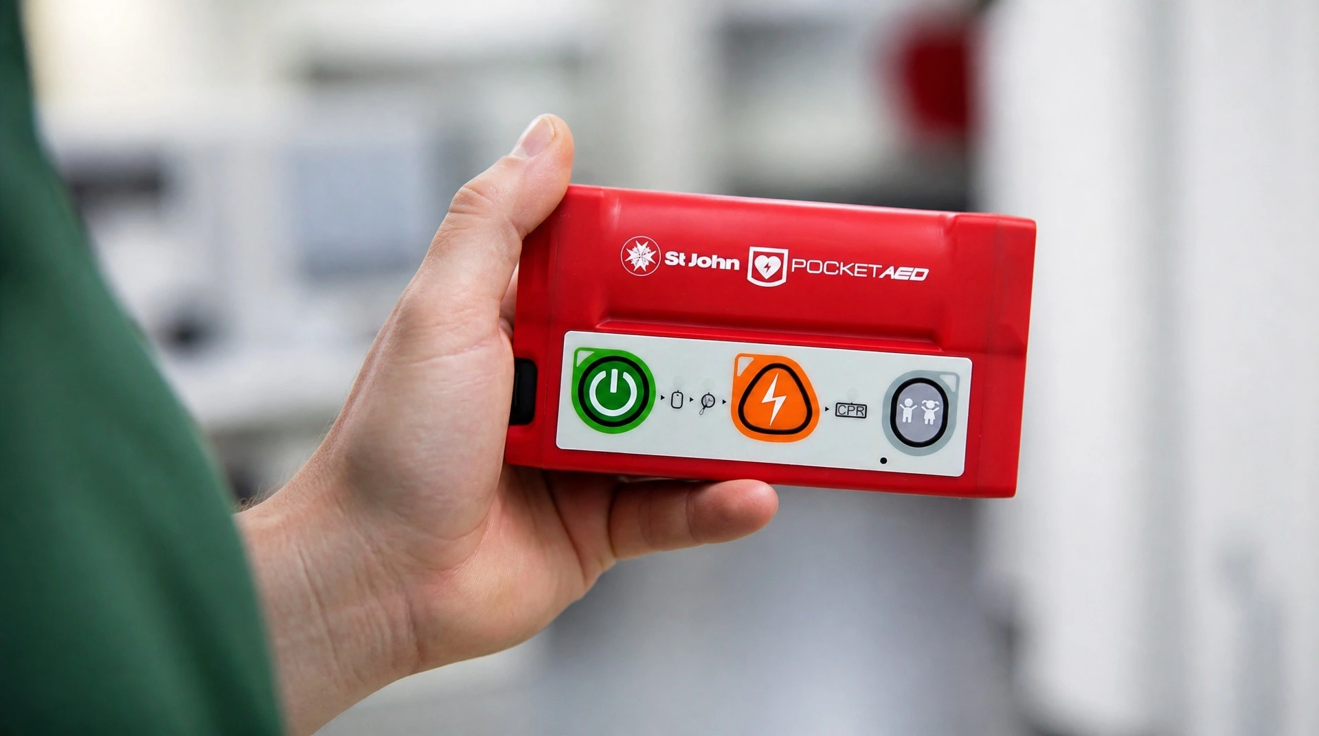 Hand holding the red St John Pocket AED, a pocket defibrillator, showing the power, shock, CPR, and child mode icon sequence on the device.